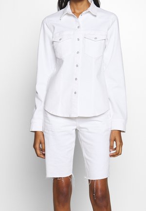 Woman wearing white denim button-up shirt and matching knee-length shorts with frayed hems, standing against a light background.