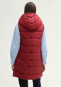 Rote Steppweste mit einer hohen, gesteppten Kapuze, die ein Rautenmuster aufweist und einen langen Schnitt hat. Getragen über einem hellblauen Hemd mit glatter Textur.