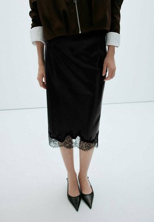 Persona que lleva una falda midi negra con ribete de encaje, tacones negros de punta afilada y una chaqueta marrón con puños blancos y cremallera frontal.