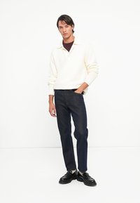 Pulover alb texturat cu guler, asortat cu blugi negri și pantofi negri. Modelul stă cu o mână în buzunarul pantalonilor.