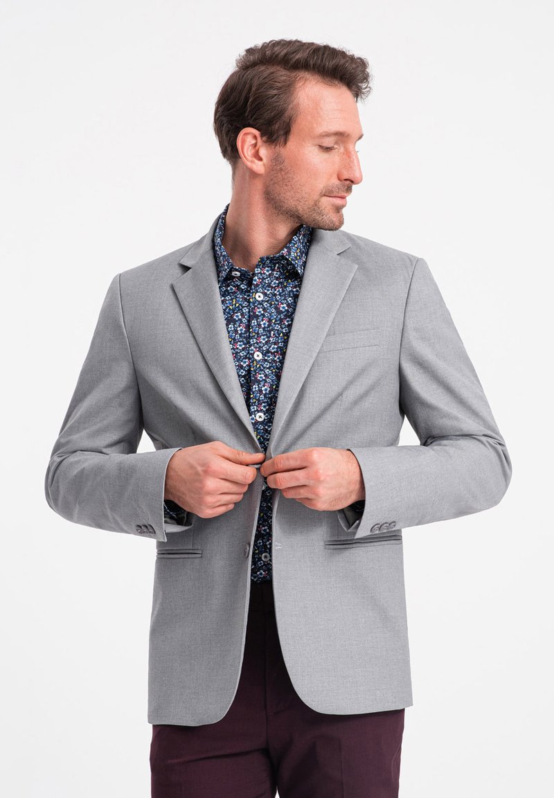 Vaaleanharmaa bleiseri, jossa on viistot läpät, etutaskut ja nappisuljenta, kerroksittain tumman kukkakuosisen paidan ja tummahaalareiden päällä.