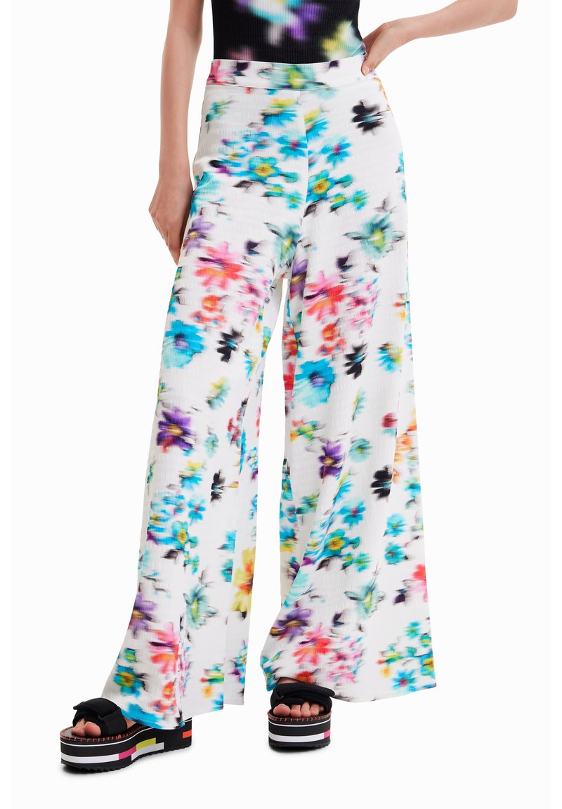 Femme portant un pantalon blanc taille haute à jambes larges avec un imprimé floral coloré, associé à des sandales à plateforme noires ornées de nœuds.