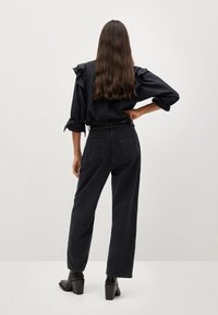 Mujer con cabello largo castaño, vistiendo una blusa negra con volantes, jeans negros de tiro alto y botines negros de tacón, de pie con una mano en la cadera.