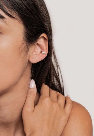 Srebrna ucha z motywem kwiatowym, z małym kryształowym akcentem. Modelka ma jasną skórę, a jej dłoń opiera się na ramieniu.