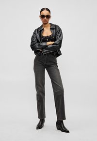 Γυναίκα με μαλλιά αναρριχημένα προς τα πίσω, φοράει μαύρα γυαλιά ηλίου, μαύρο δερμάτινο μπουφάν, μαύρο Τ-shirt με τύπωμα, σκούρο τζιν και μαύρα μποτάκια μέχρι τον αστράγαλο, στέκεται με σταυρωμένα τα χέρια.