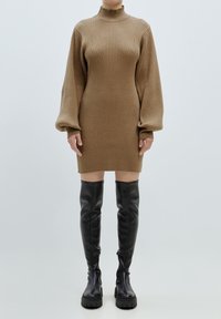 Strickkleid in Hellbraun mit hohem Kragen und gerippter Textur; kombiniert mit knielangen schwarzen Lederstiefeln mit kräftigen Sohlen.