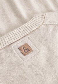 Hellbeige gestrickter Pullover mit einem gerippten Ausschnitt. Verfügt über ein quadratisches Lederpatch mit einem geprägten Und-Zeichen. Weiche Textur.