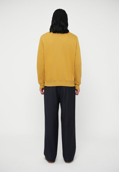 Μουσταρδί φούτερ με απλό σχέδιο, εφαρμοστά μανίκια και χαλαρή εφαρμογή, συνδυασμένο με σκούρα μπλε παντελόνι με ρίγες και φαρδιά πόδια.