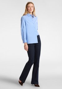 Camicia azzurra chiara con righe verticali, colletto, abbinata a jeans svasati in denim scuro e tacchi a punta marroni. Texture liscia.