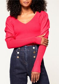 Donna con capelli ricci che indossa un caldo top rosa brillante a maniche lunghe a coste con spalle a sbuffo e jeans scuri a vita alta con bottoni dorati.