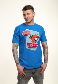 Modrá bavlněná tričko s grafikou usměvavé oranžové krávy, doplněné textem a barevnými akcenty, nošené s vybledlými šedými džínami.