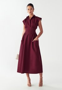 Abito borgogna con maniche corte a cappuccio, colletto chiuso da bottoni, cucitura in vita e due grandi tasche laterali. Tessuto liscio, silhouette A-line fluida.