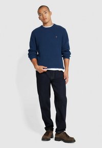 Granatowy sweter prążkowany z okrągłym dekoltem, noszony na białej koszuli, zestawiony z ciemnymi dżinsami i brązowymi butami, z subtelnym detalem logo.