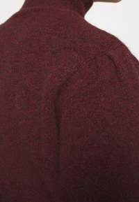 Pull en tricot bordeaux avec un col montant et un tissu texturé et doux. Présente des manches légèrement bouffantes et une silhouette ajustée.