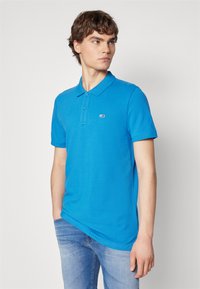 Polo shirt blu in cotone con maniche corte, chiusura a tre bottoni e un piccolo logo sul petto sinistro. Indossato con jeans azzurri chiari.