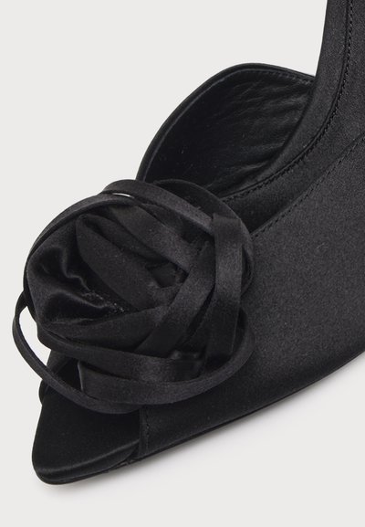 Chaussure noire à talon haut avec bout ouvert et fleur décorative en ruban de satin noir sur le dessus, sur un fond clair uni.