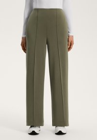 Ohlapne, olivno zelene hlače širokega kroja, izdelane iz mehkega materiala; vsebujejo vertikalne šive, poudarke na robu in so kombinirane z belim obutvijo.