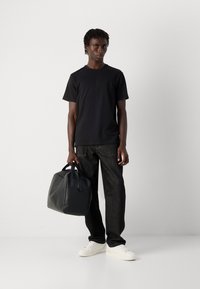 Man in een zwart T-shirt, donkere jeans en witte sneakers die tegen een witte muur staat en een zwart leren weekendtas bij de handvatten vasthoudt.