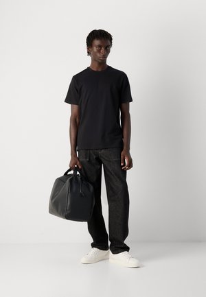 Man in een zwart T-shirt, donkere jeans en witte sneakers die tegen een witte muur staat en een zwart leren weekendtas bij de handvatten vasthoudt.