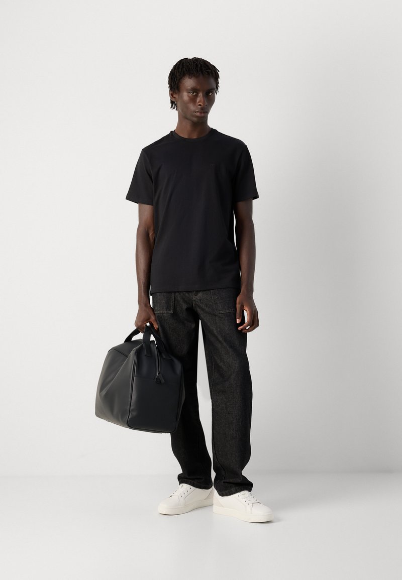 Man in een zwart T-shirt, donkere jeans en witte sneakers die tegen een witte muur staat en een zwart leren weekendtas bij de handvatten vasthoudt.