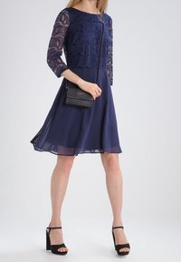 Žena ve tmavě modrých krajkových šatech s průhlednými rukávy, držící malou černou crossbody kabelku a černé sandály na vysokém podpatku.