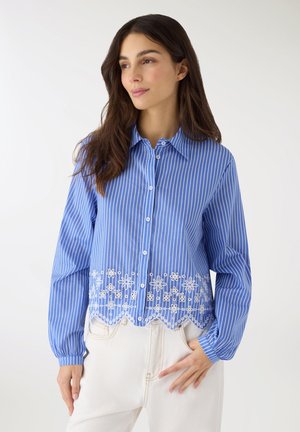 Giovane donna che indossa una camicetta a righe blu e bianche con ricami floreali sull'orlo, abbinata a pantaloni bianchi, posando su sfondo bianco.