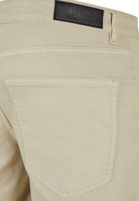 Jeans de mezclilla de algodón color beige con una etiqueta de cuero que dice "URBAN CLASSICS" y un único bolsillo trasero con costuras decorativas.