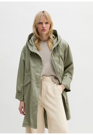 Blonde vrouw met een lichtgroene jas met capuchon, beige gebreide trui en crèmekleurige broek met brede pijpen, staand met hand in de zak tegen een witte achtergrond.