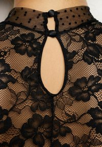 Svart spetsblus med blommönster, genomskinlig prickig krage, öppen rygg, knäppt med svarta knappar, som framhäver intrikata texturer.