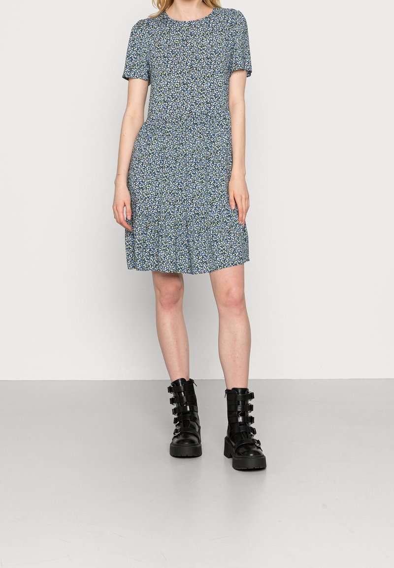 Femme portant une robe bleue à manches courtes à fleurs et des bottes de combat noires à boucle, debout sur un sol gris clair contre un mur blanc.