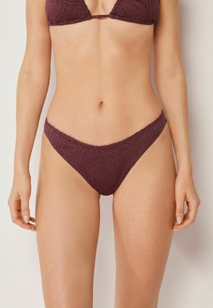 Kvinde iført bordeaux bikinitrusser med paisleymønster og matchende top, stående mod en ensartet lys baggrund.