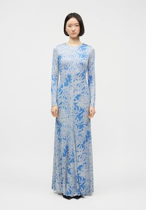 Moteris trumpais juodais plaukais, vilkinti ilgomis rankovėmis, grindų ilgio mėlyną suknelę su baltų lapų raštais, stovinti prieš baltą foną.