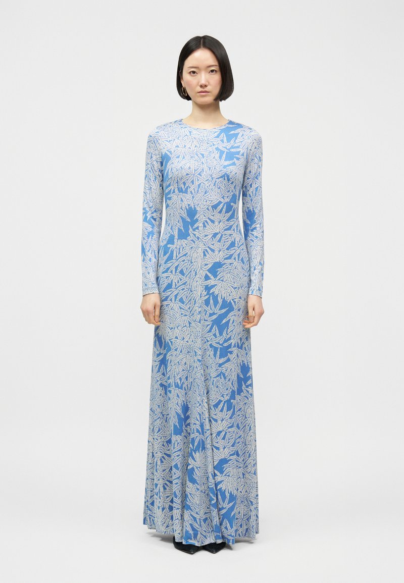 Γυναίκα με κοντά μαύρα μαλλιά, φοράει μπλε φόρεμα μακριάς μανίκας μέχρι το πάτωμα με άσπρα σχέδια φύλλων, στεκόμενη μπροστά σε λευκό φόντο.