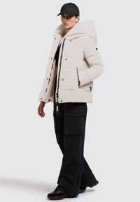 Kurtka puchowa w jasnym beżu z dużym kapturem, zapięciem na zamek i bocznymi kieszeniami; zestawiona z czarnymi spodniami cargo i czarnymi trampkami.
