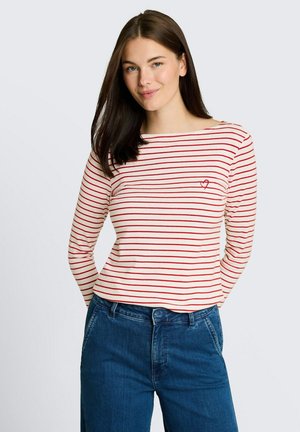 Jeune femme aux cheveux lisses et foncés portant une chemise blanche à manches longues avec des rayures rouges et un petit cœur, associée à un jean bleu.