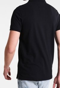 Černé polo tričko s krátkými rukávy, přiléhavý střih a boční rozparky. Tkanina se zdá být hladká, bez viditelných vzorů nebo ozdob.