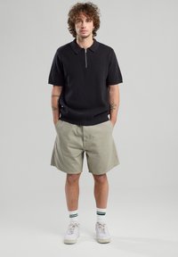 Black short-sleeve polo shirt with a zipper, paired with light gray shorts. White sneakers with green striped socks complete the outfit.