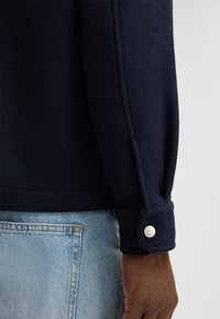 Manche de veste en polaire bleu marine avec un poignet à boutons, texture lisse et détails de couture arrondis, associée à un jean en denim bleu clair.