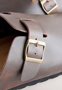 Braune Ledersandalen mit einem breiten Riemen, Messing-Schnallenbeschlägen und einer strukturierten Fußbettoberfläche. Die Ränder sind leicht abgerundet und die Sohle ist dunkel.