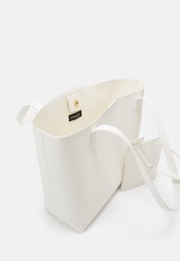 Sac fourre-tout en cuir blanc avec de longues poignées, ouverture supérieure, fermeture à pression et pochette plate attachée à l'intérieur.