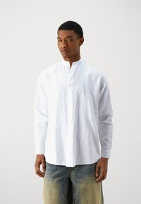 Camicia bianca a maniche lunghe con colletto alto, frontale con bottoni, realizzata in un tessuto leggero, abbinata a jeans blu sbiaditi.