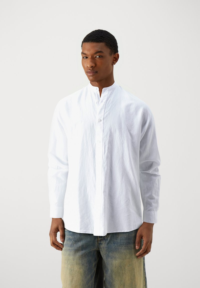 Camicia bianca a maniche lunghe con colletto alto, frontale con bottoni, realizzata in un tessuto leggero, abbinata a jeans blu sbiaditi.