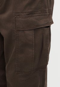 Gros plan sur la jambe d'un pantalon cargo marron avec une grande poche à rabat et des coutures visibles sur le tissu.