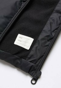 Vnitřek černé bundy se strukturovanou fleecovou podšívkou a bílou cedulkou na jméno a telefonní číslo, s viditelným zipem a klouzákem.