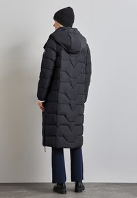 Svart uppblåsbar jacka med lång silhuett, huva och chevron-mönster. Har elastiska ärmslut och dragsko i nederkant för justerbar passform.