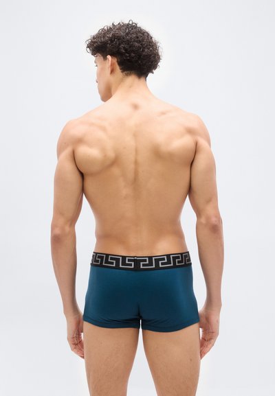 Νεαρός άνδρας με σγουρά μαλλιά στέκεται με τη πλάτη στραμμένη, δείχνοντας τον μυώδη του κορμό, φορώντας μπλε μποξεράκια με μαύρη ζώνη μέσης και άσπρο γεωμετρικό σχέδιο.