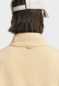 Veste en polaire beige avec une finition texturée, dotée d'un col haut et d'un petit détail en boucle à l'arrière. Casquette avec sangle ajustable visible.