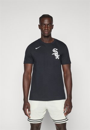 Camiseta de algodón negra de manga corta con el logo blanco de "Sox" y el swoosh de Nike. Llevada con pantalones cortos perforados de color claro y rayas negras.