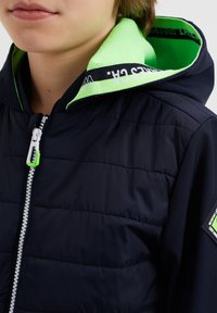 Námořnická vyteplená bunda s černou a zelenou podšívkou kapuce, která má přední zip a logo na rukávu. Viditelné detaily strukturované látky.