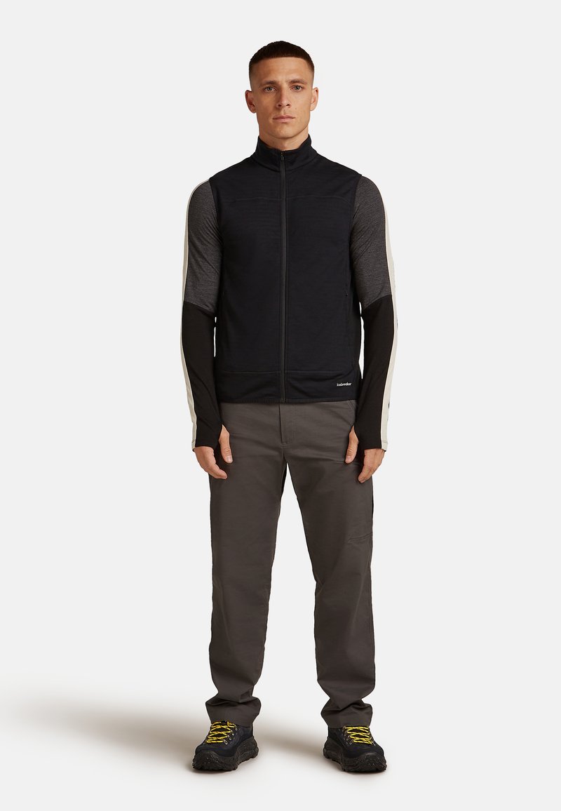 Un giovane uomo sta in piedi rivolto in avanti, indossando un gilet nero senza maniche con zip su una maglietta a maniche lunghe nera e grigia, pantaloni grigi e scarpe nere con lacci gialli.